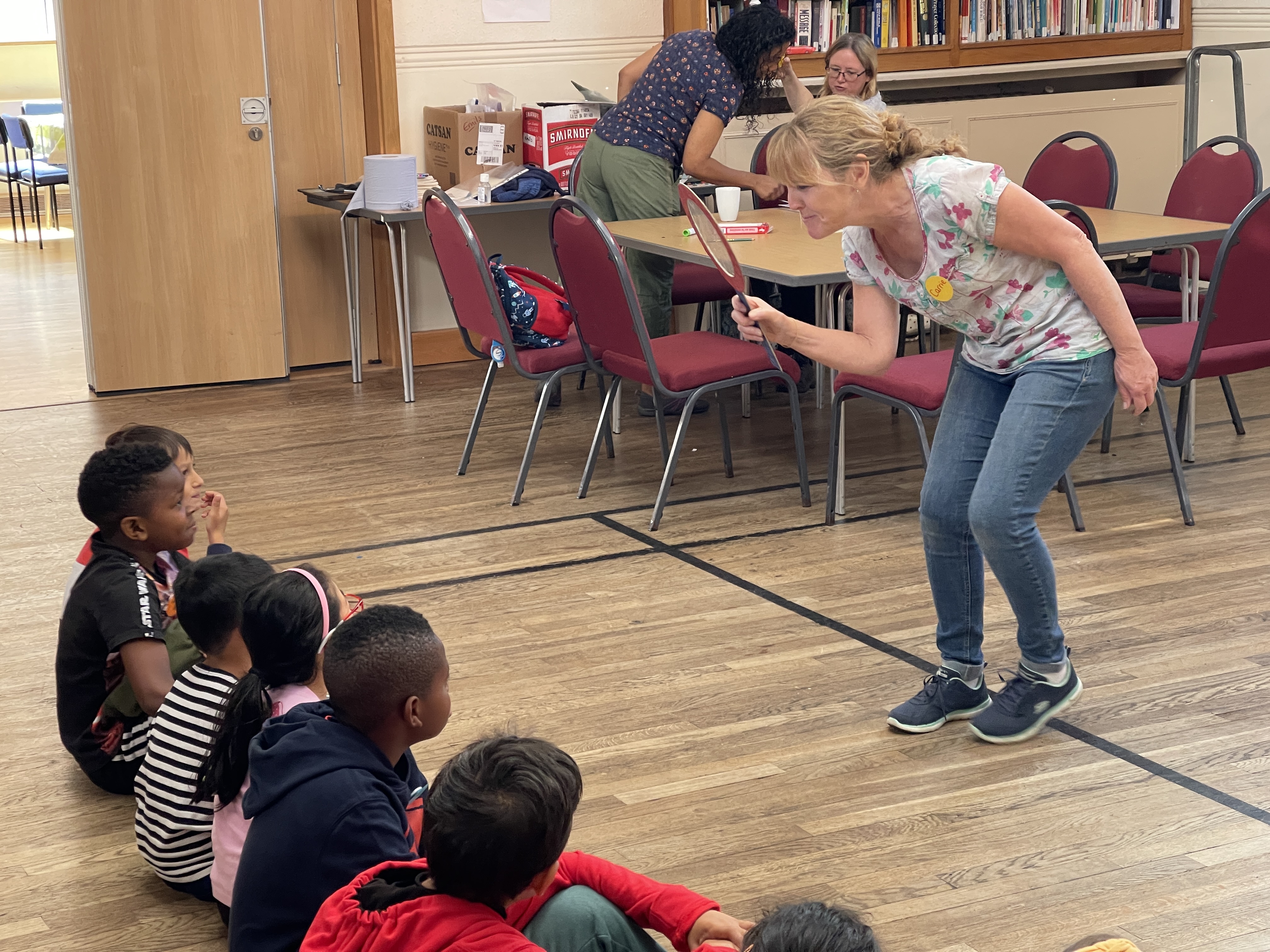 A group of children sitting on floor while an adult leader looks at them through a large magnifying glass